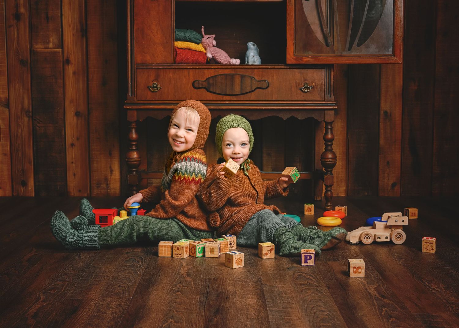 Two children in matching brown sweaters play with wooden blocks on hardwood floor.