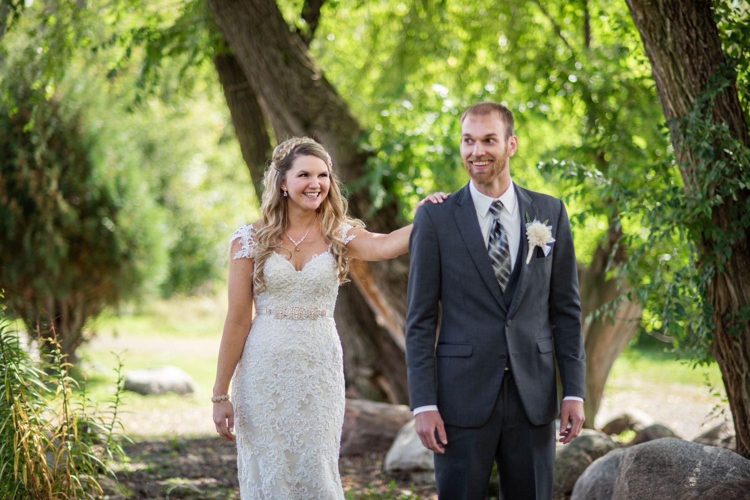 A romantic wedding couple stands together on a path surrounded by lush green trees and natural landscaping.