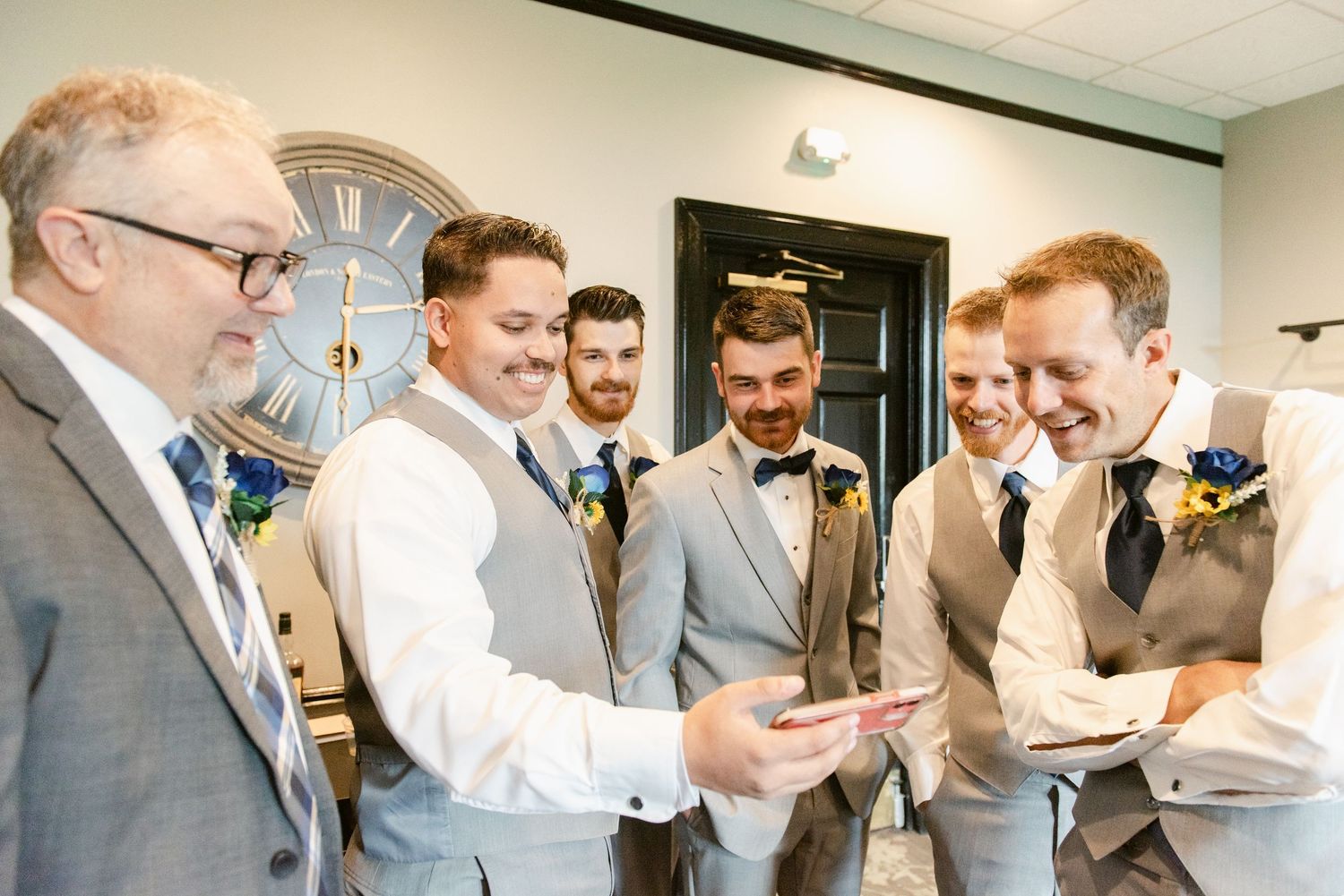 A group of groomsmen in gray and white attire gather in a room to look at something on a phone.