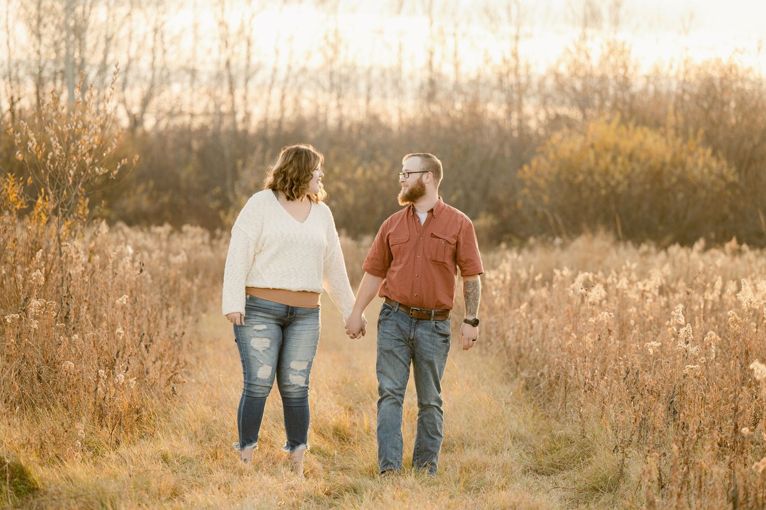 Two figures holding hands walk together through a golden field at sunset in a romantic outdoor setting. St cloud area engagement photos.