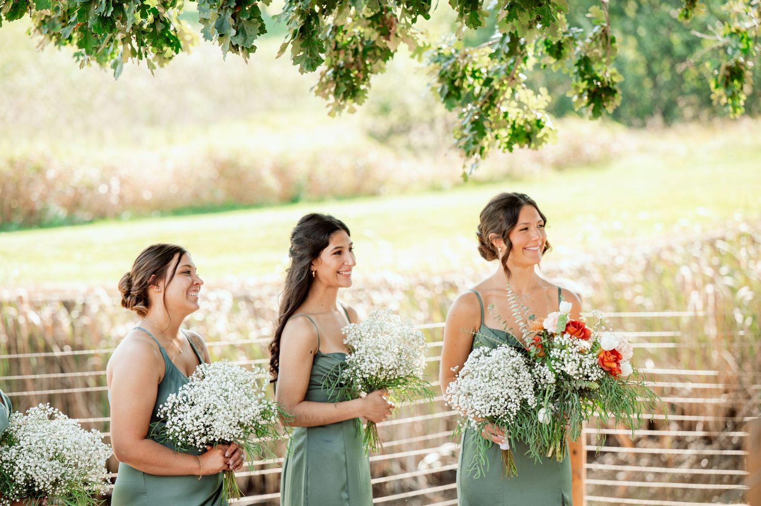 Three bridesmaids in sage green dresses hold flower bouquets while standing outdoors.