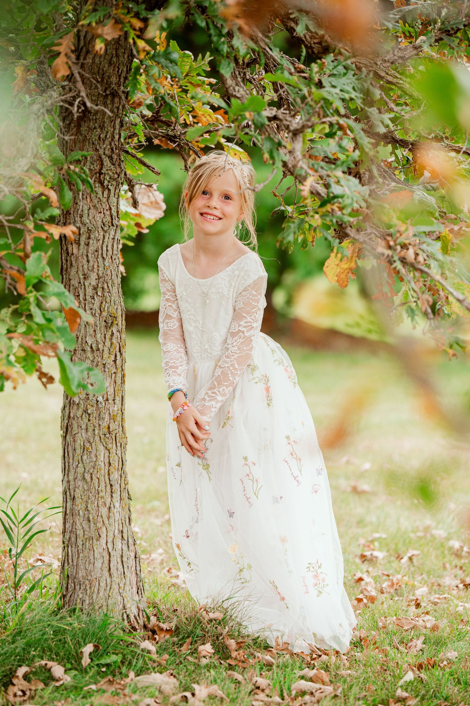 Young child in long white lace dress standing by tree trunk in autumn garden setting with falling leaves.