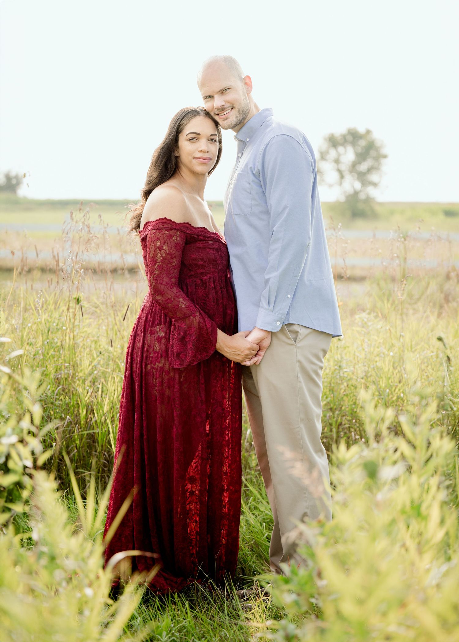 A couple poses together in tall grass, with burgundy lace dress and light blue button-down shirt.