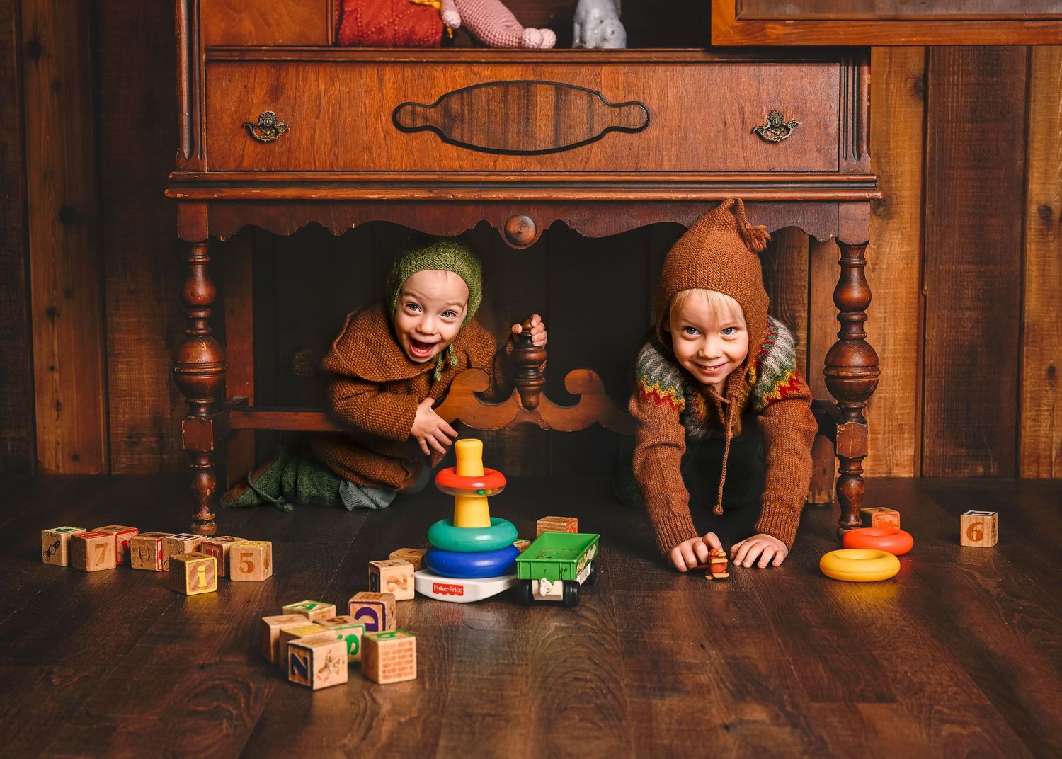 Two children in brown sweaters play with colorful toys under wooden furniture.