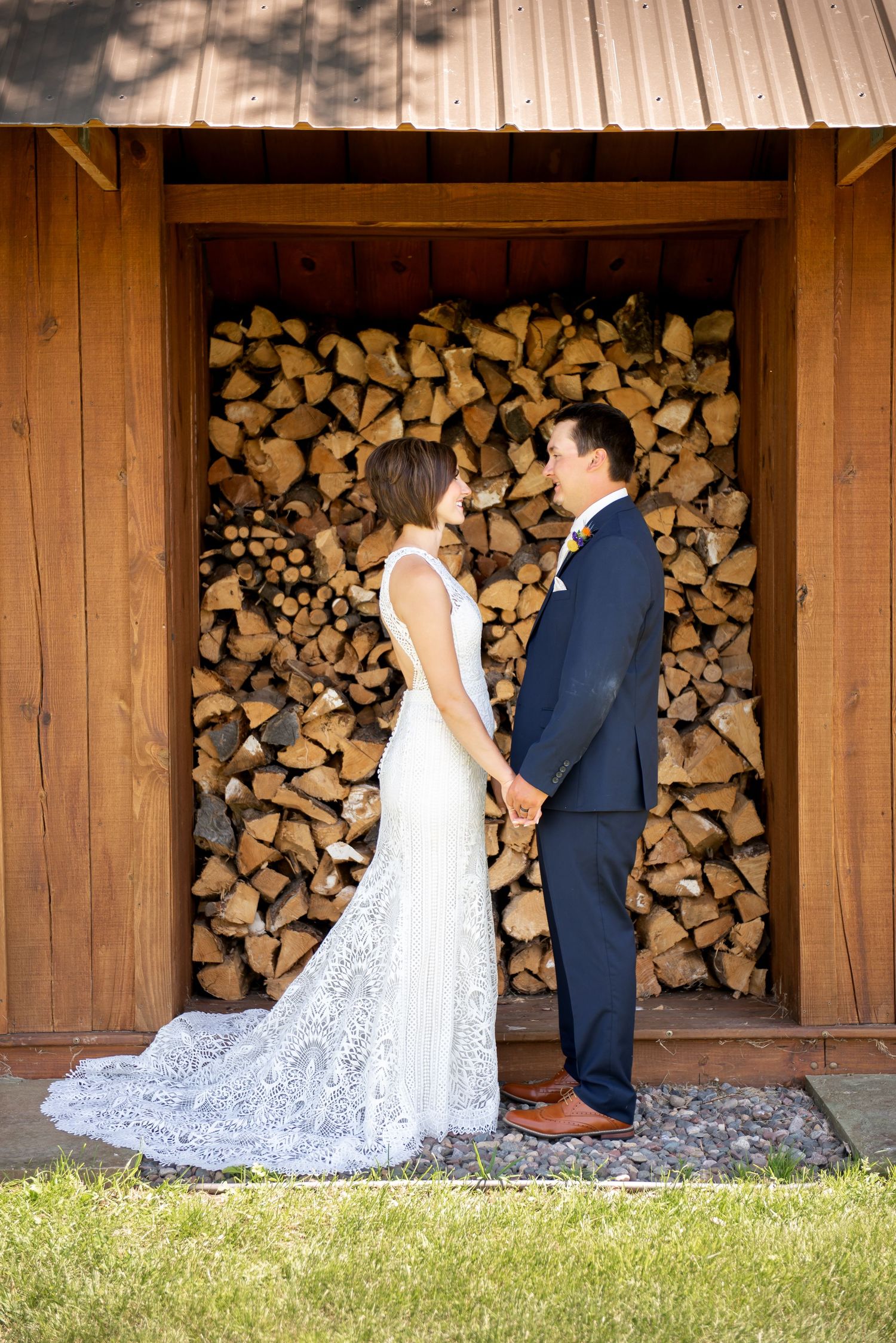 Couple in formal wedding attire pose against rustic wooden wall with stacked firewood in background.