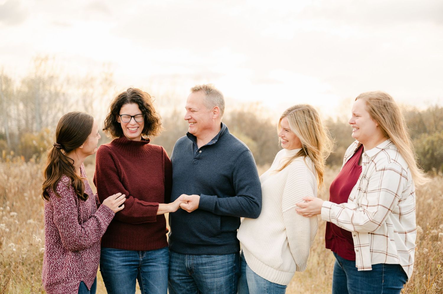 A smiling family of five wearing cozy sweaters stands together outdoors during golden hour. Princeton MN Milestone, Maternity and Newborn P