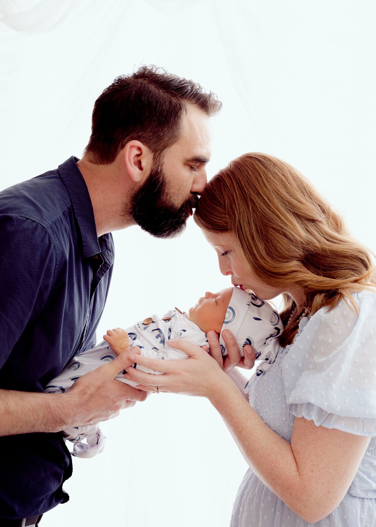Intimate family moment with parents cradling newborn baby wearing patterned outfit against white backdrop.