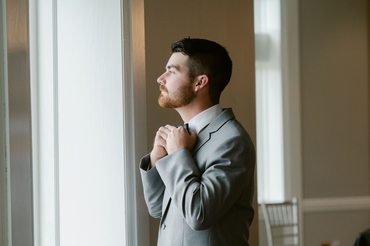 A groom in a light gray suit gazes thoughtfully out a window while adjusting his tie.