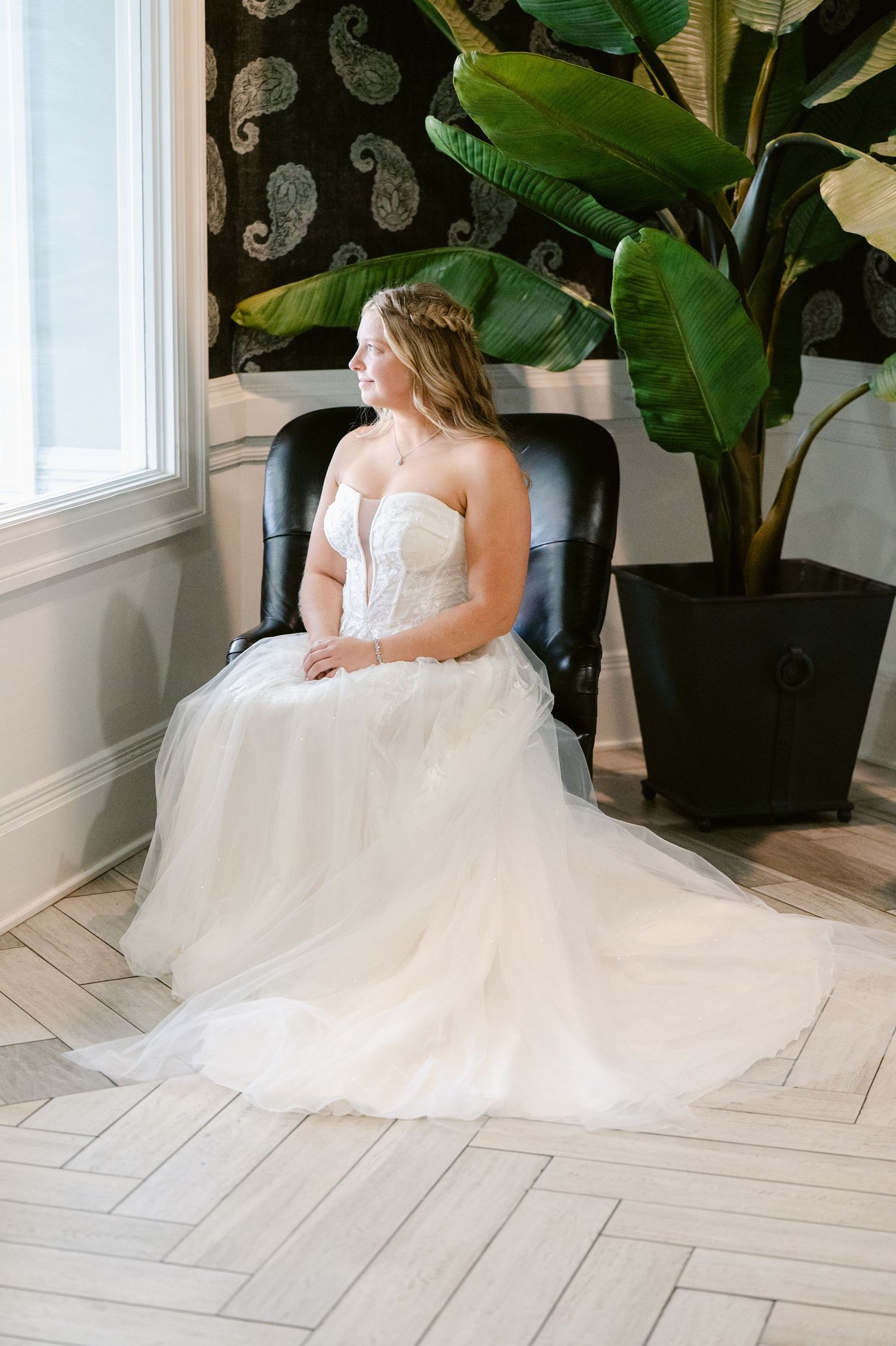 A bride in a flowing white strapless ballgown sits gracefully beside lush green tropical plants in a sunlit room.