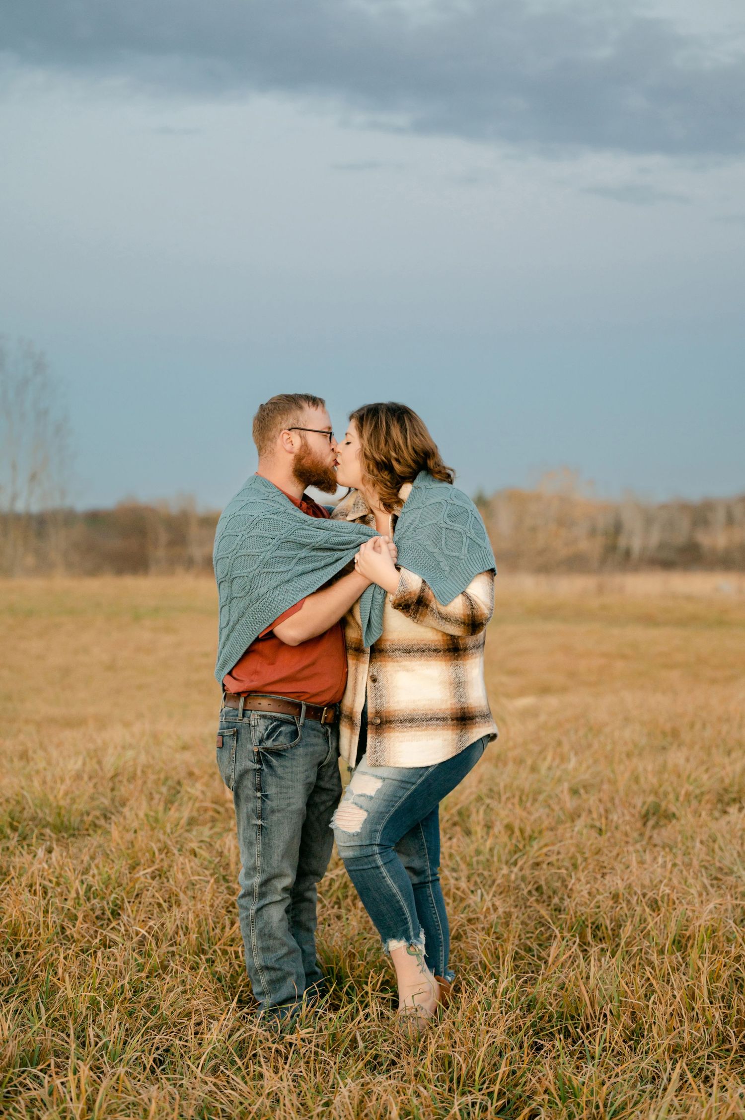 A couple shares a kiss while standing together in a golden autumn field under moody skies. St cloud area engagement photos.