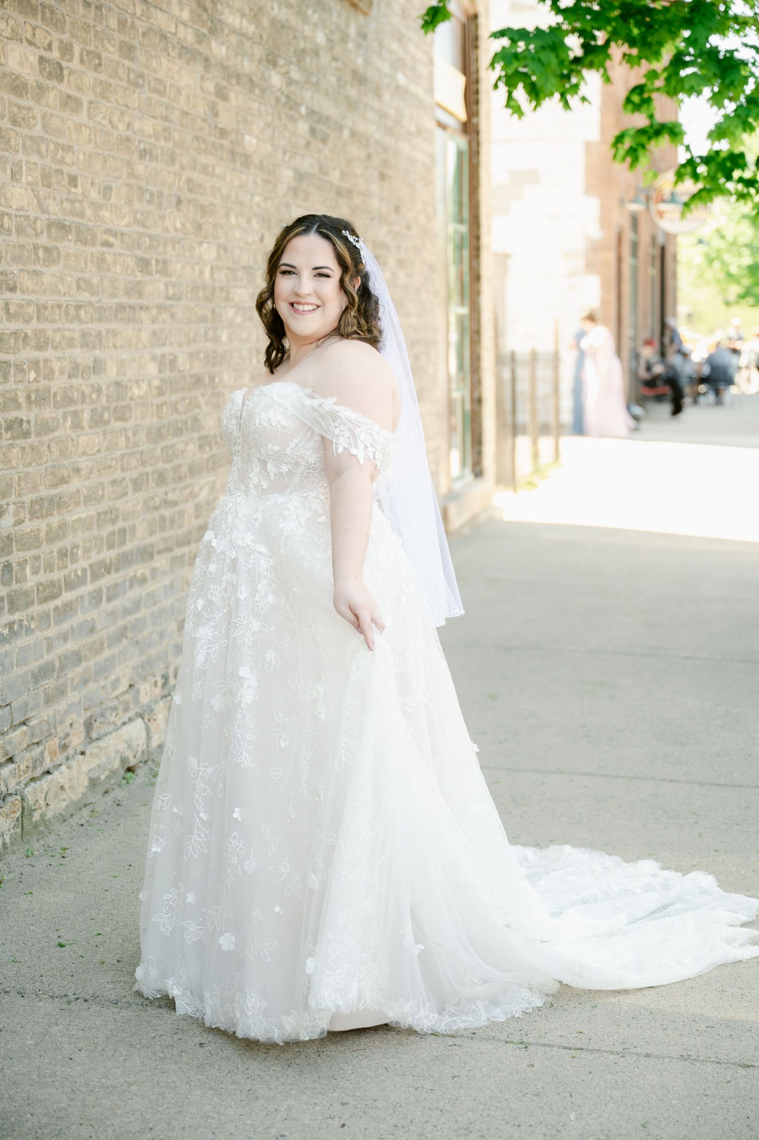 Elegant bride in lace off-shoulder wedding gown poses against brick wall on sunny day.