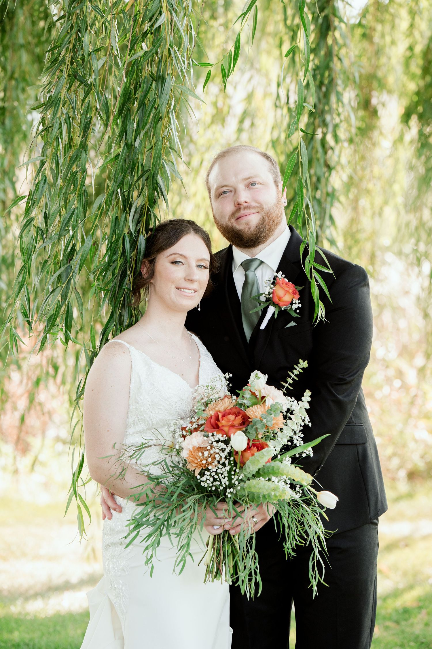 The Grands at Mulligans- Wedding Photos: couple poses under hanging willow branches with a vibrant orange and white floral bouquet.