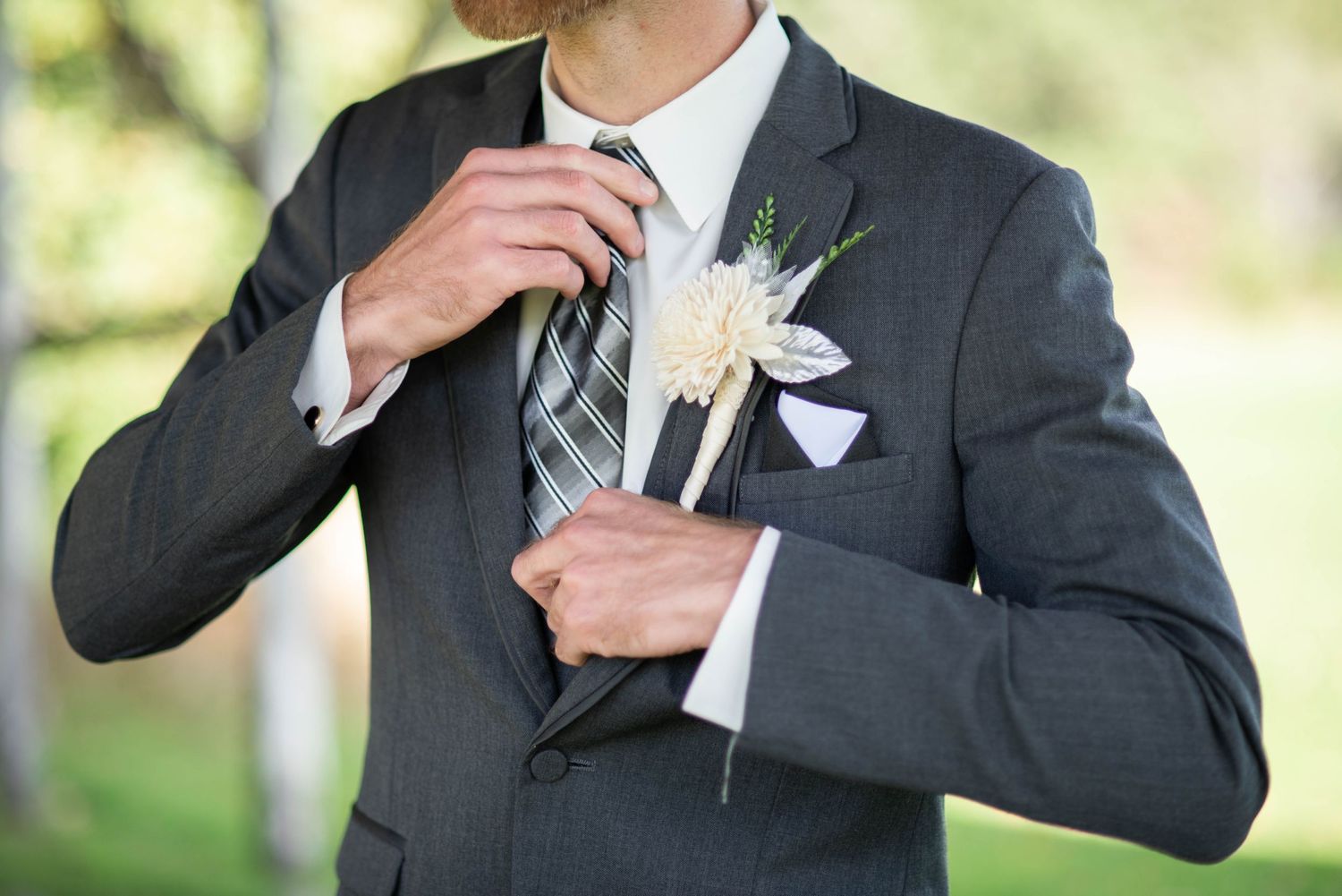 Groom adjusting his tie and jacket before wedding ceremony in natural outdoor setting.