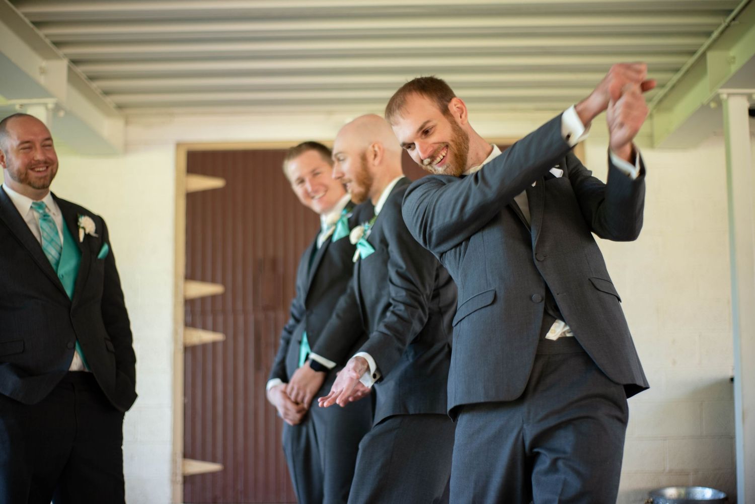 Groomsmen in black suits laugh together while standing in a row outside a building.