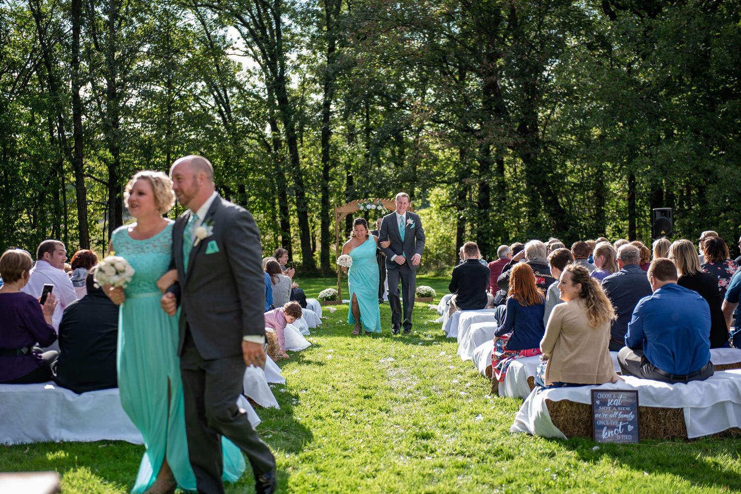 Wedding ceremony processional on sunny day with guests seated on white chairs in forest clearing. Rum River Barn Milaca Minnesota Weddings