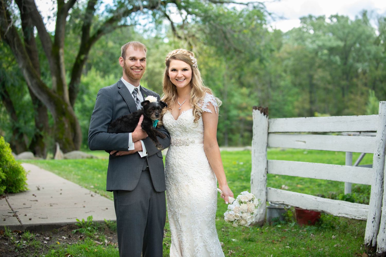 Newlyweds pose with their small dog by white fence on wedding day. Rum River Barn Milaca Minnesota Weddings