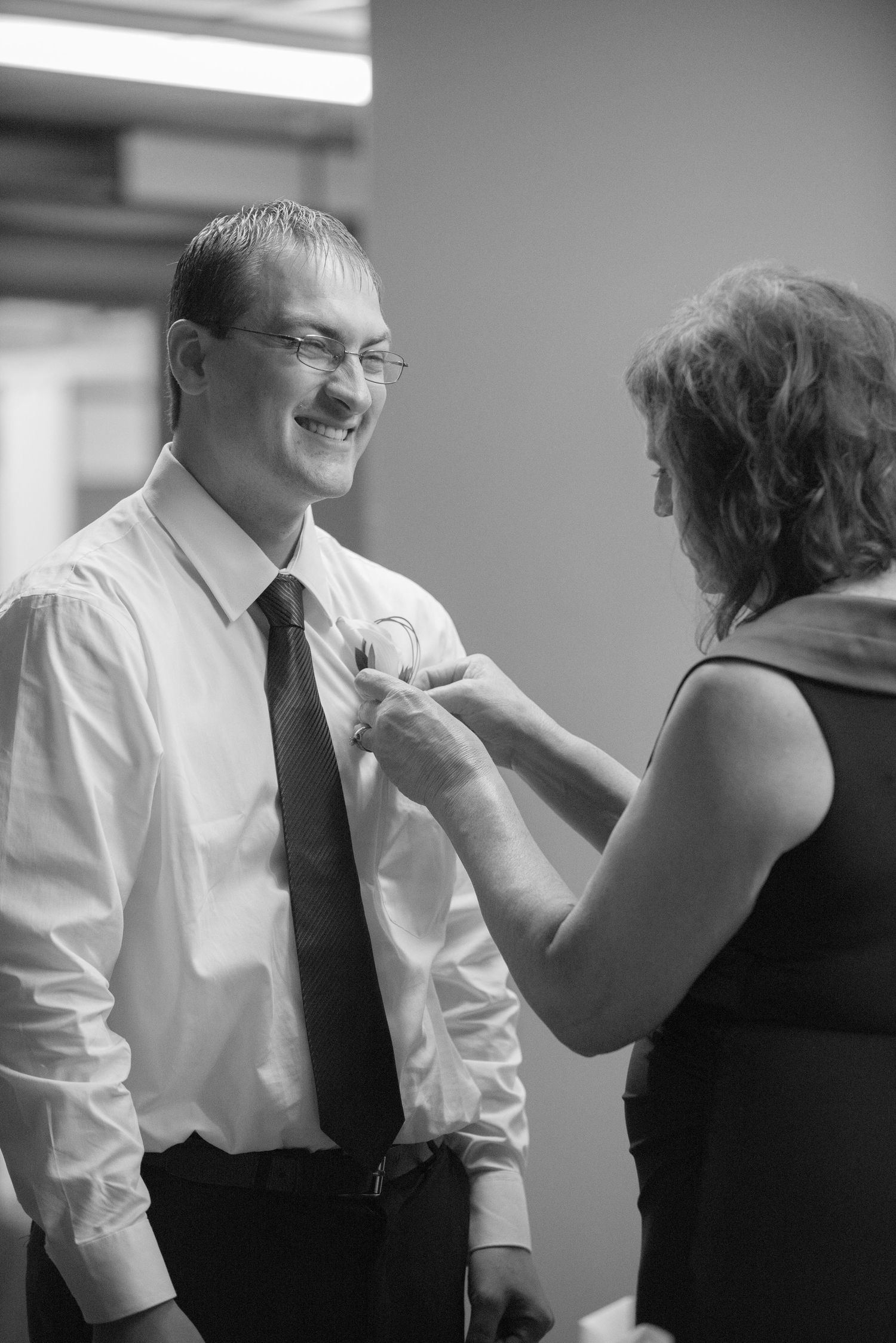 A black and white photo of someone helping to pin a boutonniere on a smiling guest at a formal event. Andover wedding at Magestic Oaks Golf