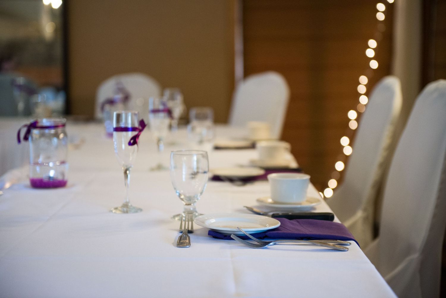 Wedding reception table setting with white linens, purple accents, and glowing string lights in background. Andover wedding at Magestic Oaks