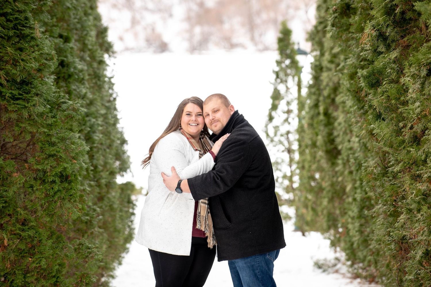 A couple embraces warmly while standing between two evergreen hedges in a snowy winter setting.