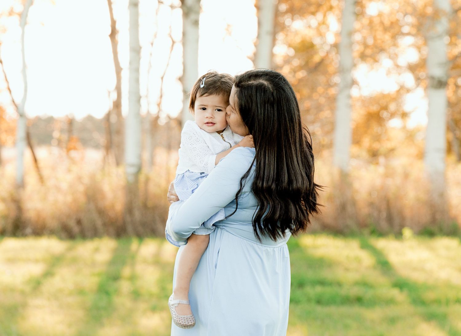 A woman in a blue dress holds a small child while standing in front of birch trees at sunset.