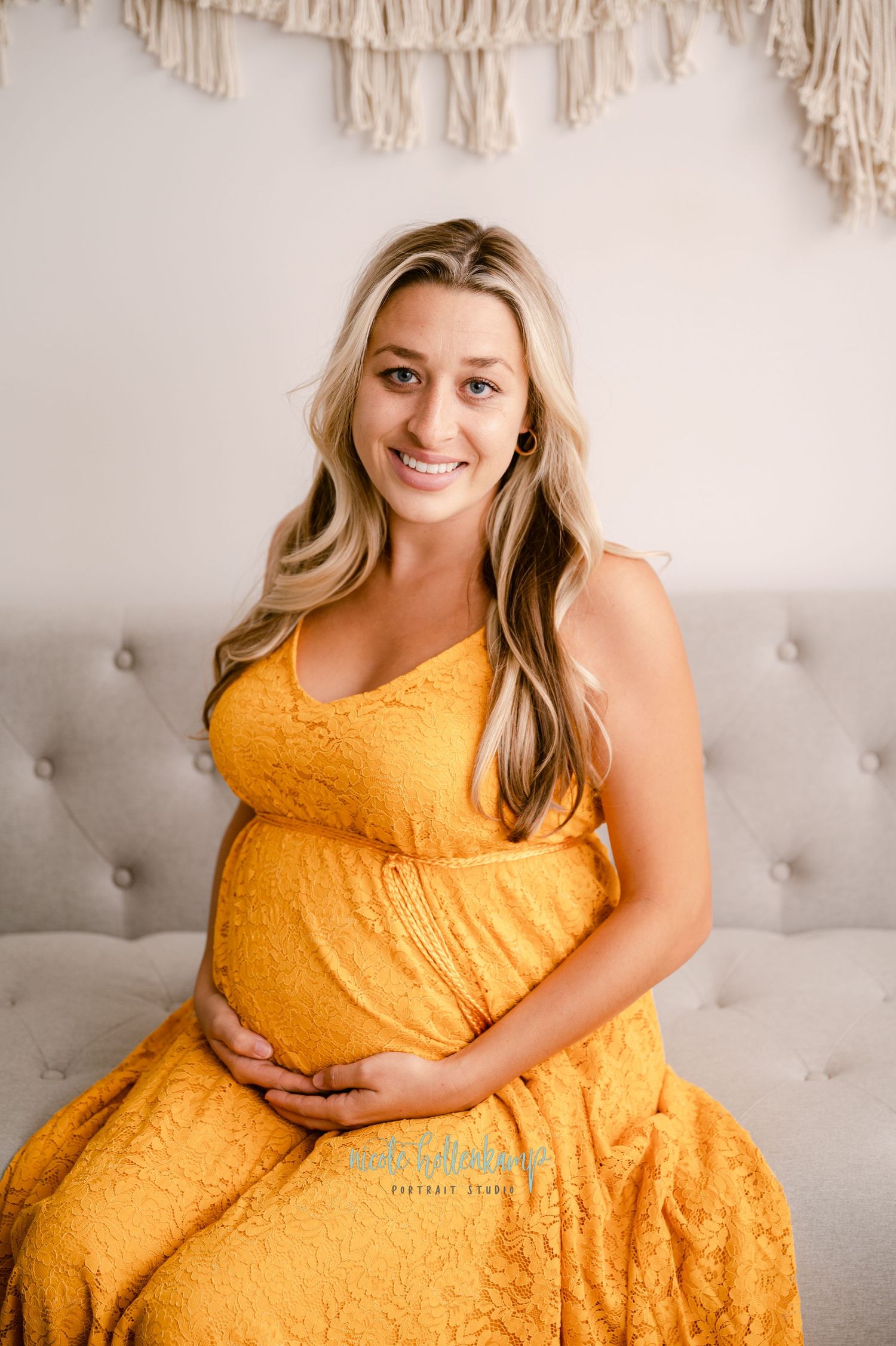 A woman in a marigold yellow lace maternity dress sits on a gray couch against a cream wall with macrame decor.