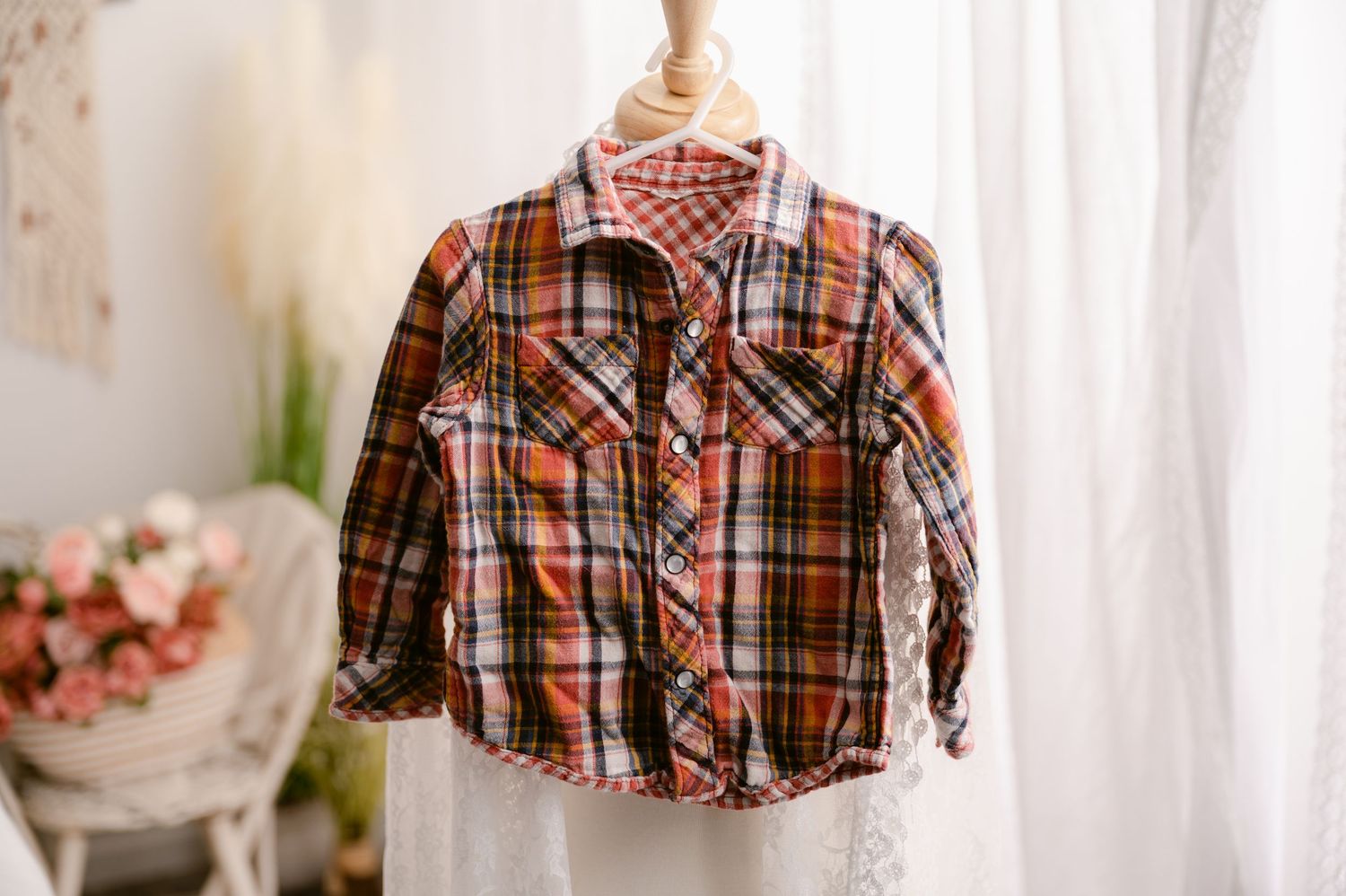 A plaid brown and orange button-down shirt hangs on a white hanger against a light-colored curtained background.