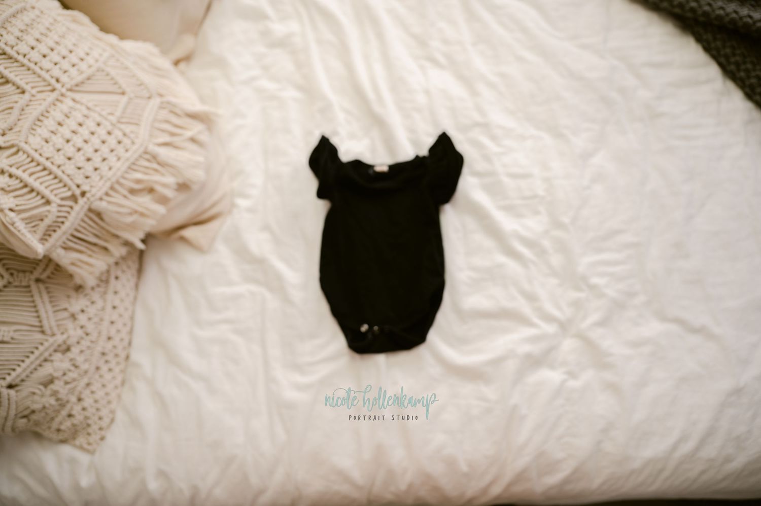 A black bodysuit or onesie garment laid flat on white bedding next to cream-colored knit fabric.