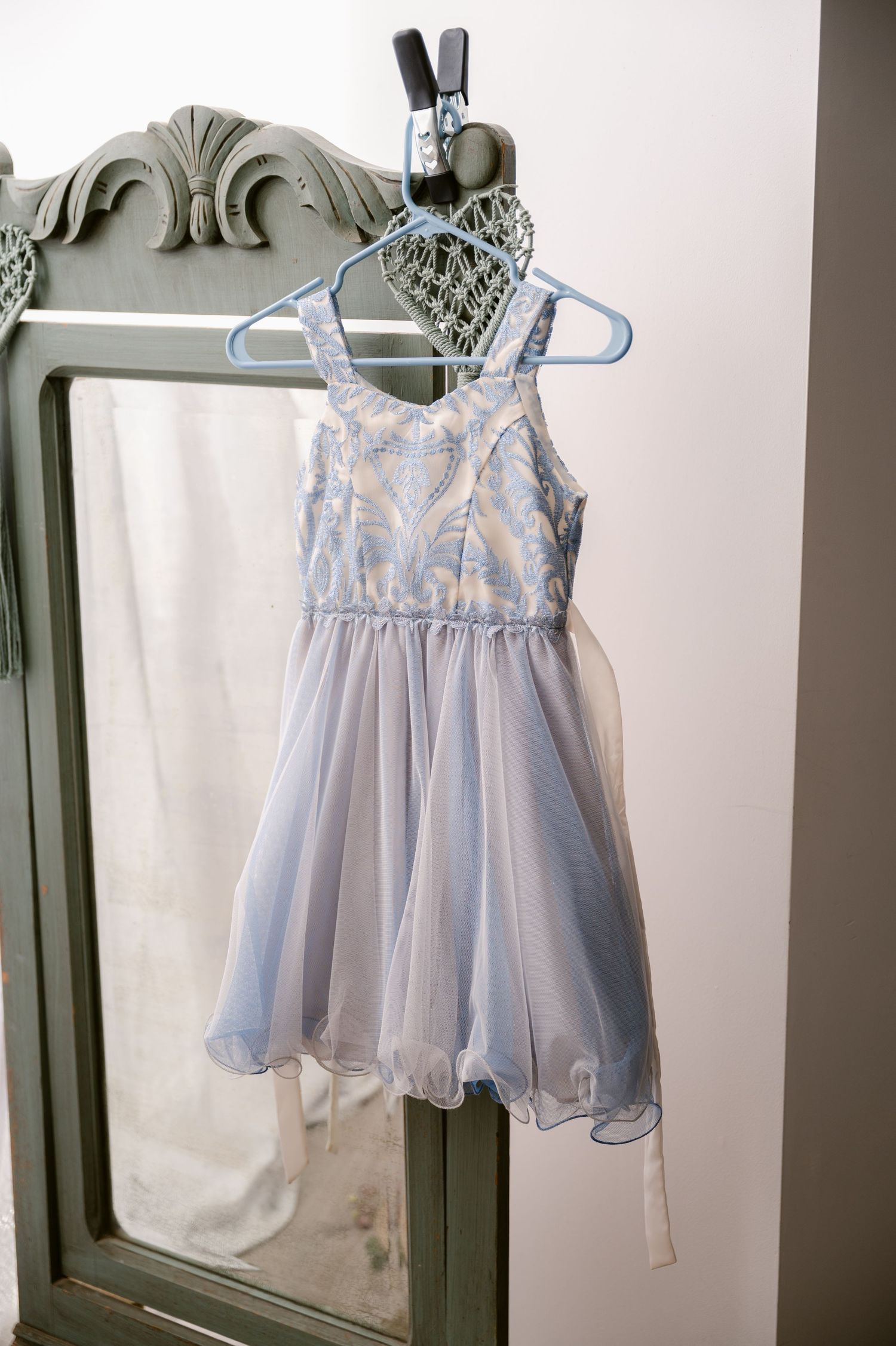 A pale blue tulle party dress with floral details hangs on a white hanger against an antique sage green armoire.