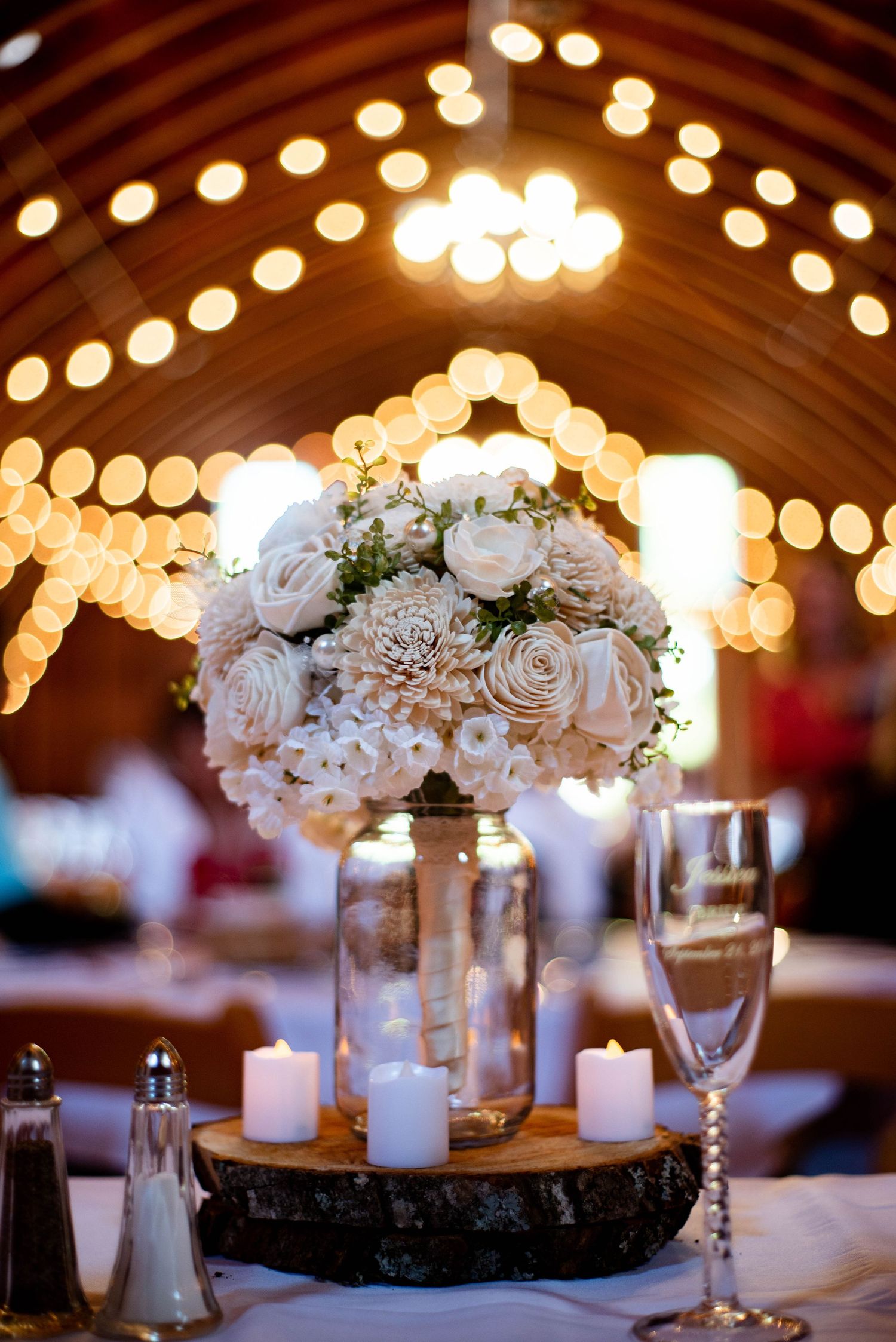 Elegant wedding reception table centerpieces with white roses and glowing string lights in rustic wooden venue.
