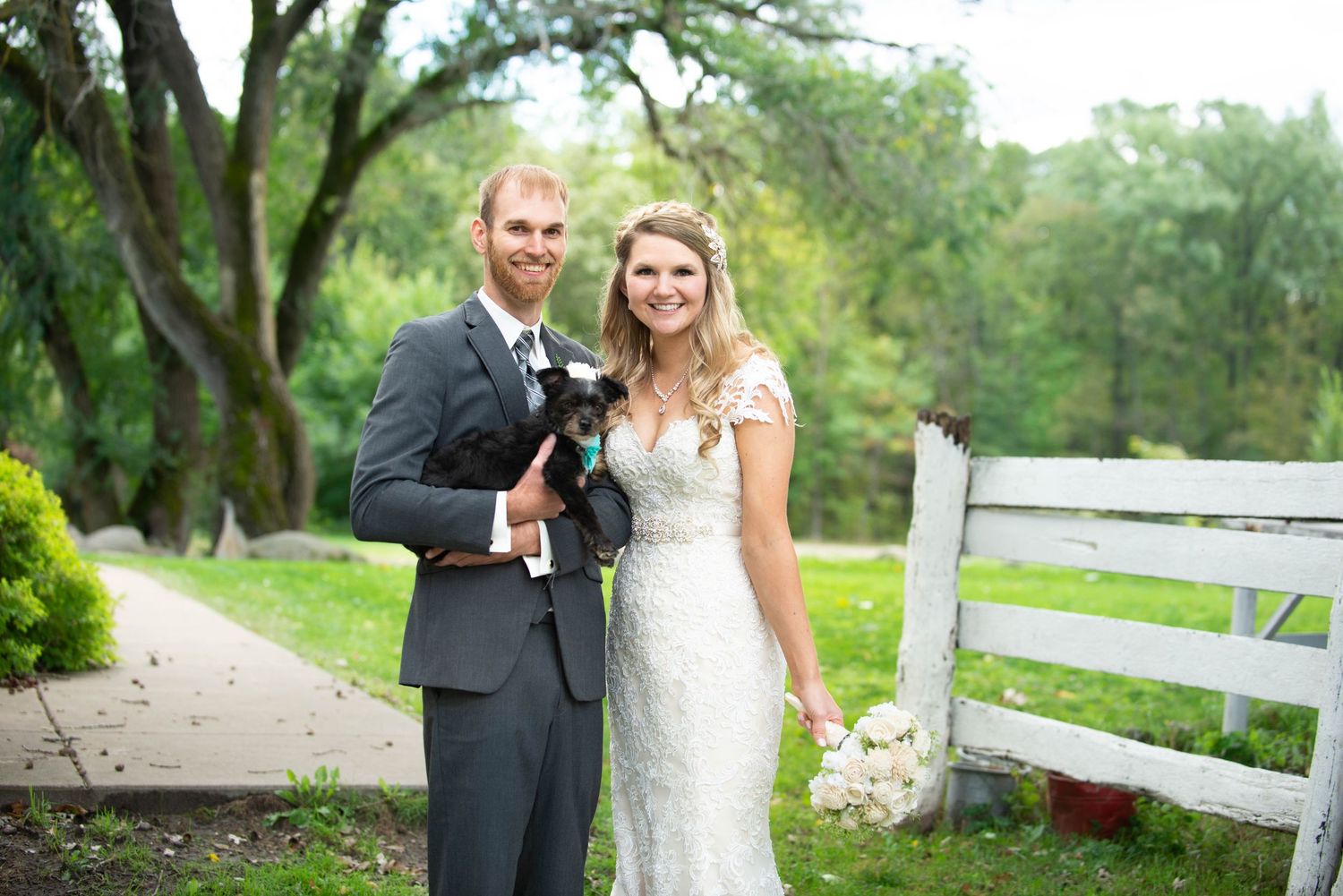 Newlyweds pose together on a path with white fencing and green trees in background. Rum River Barn Milaca Minnesota Weddings