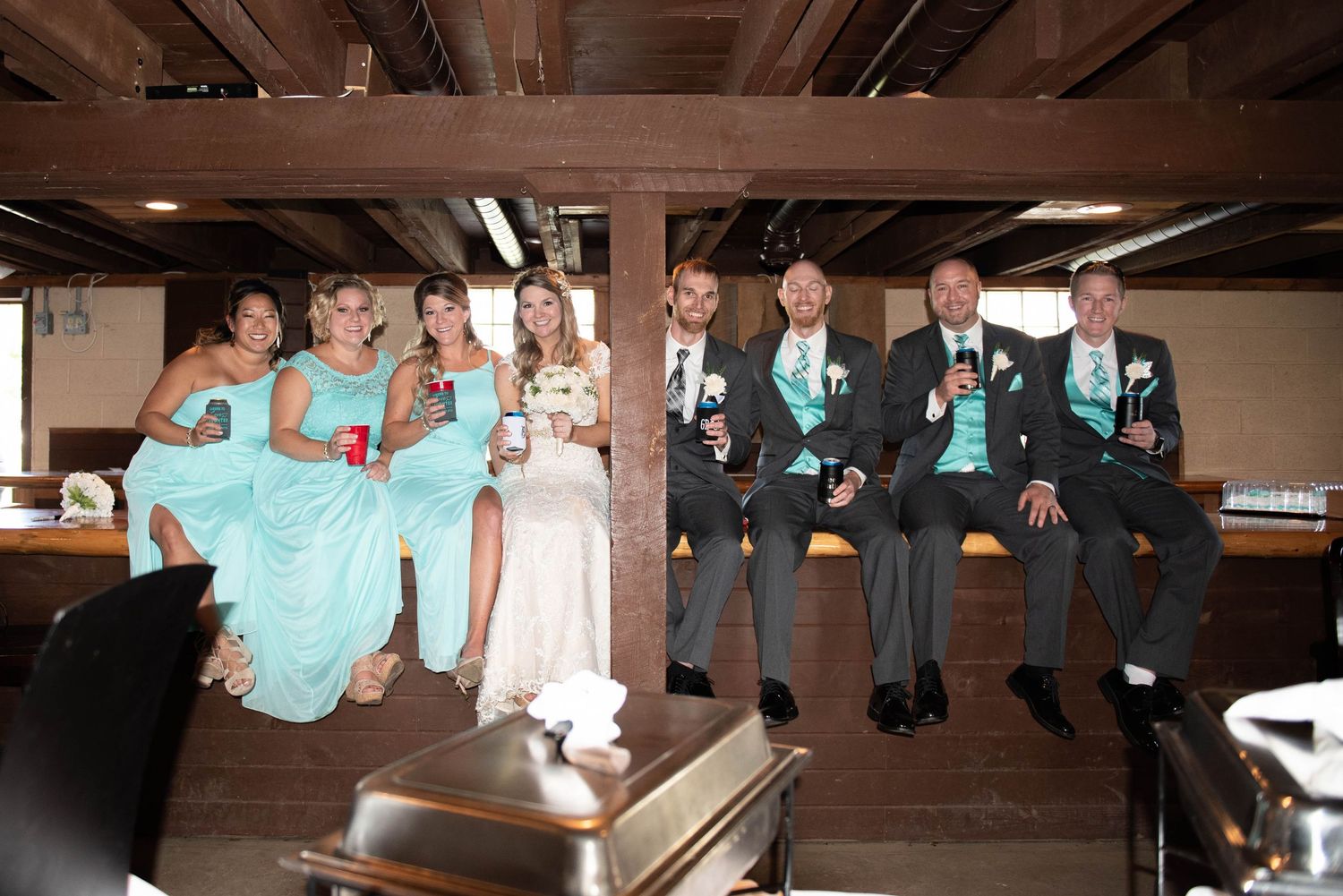 Wedding party in turquoise dresses and grey suits sitting casually on wooden beam. Rum River Barn Milaca Minnesota Weddings