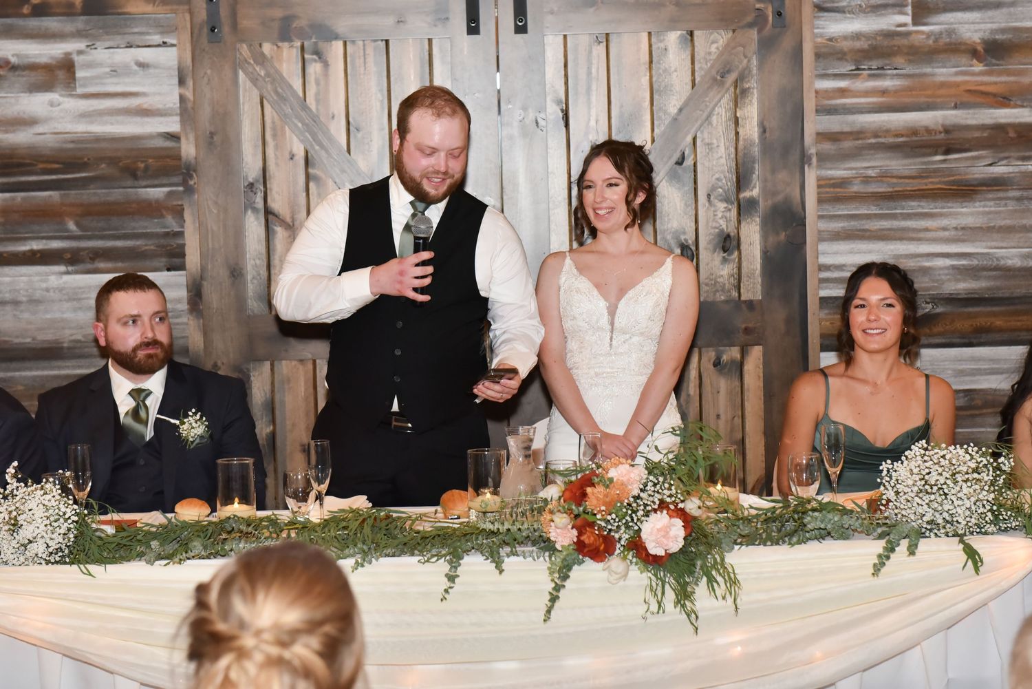 Wedding party seated at rustic wooden head table with floral decorations.