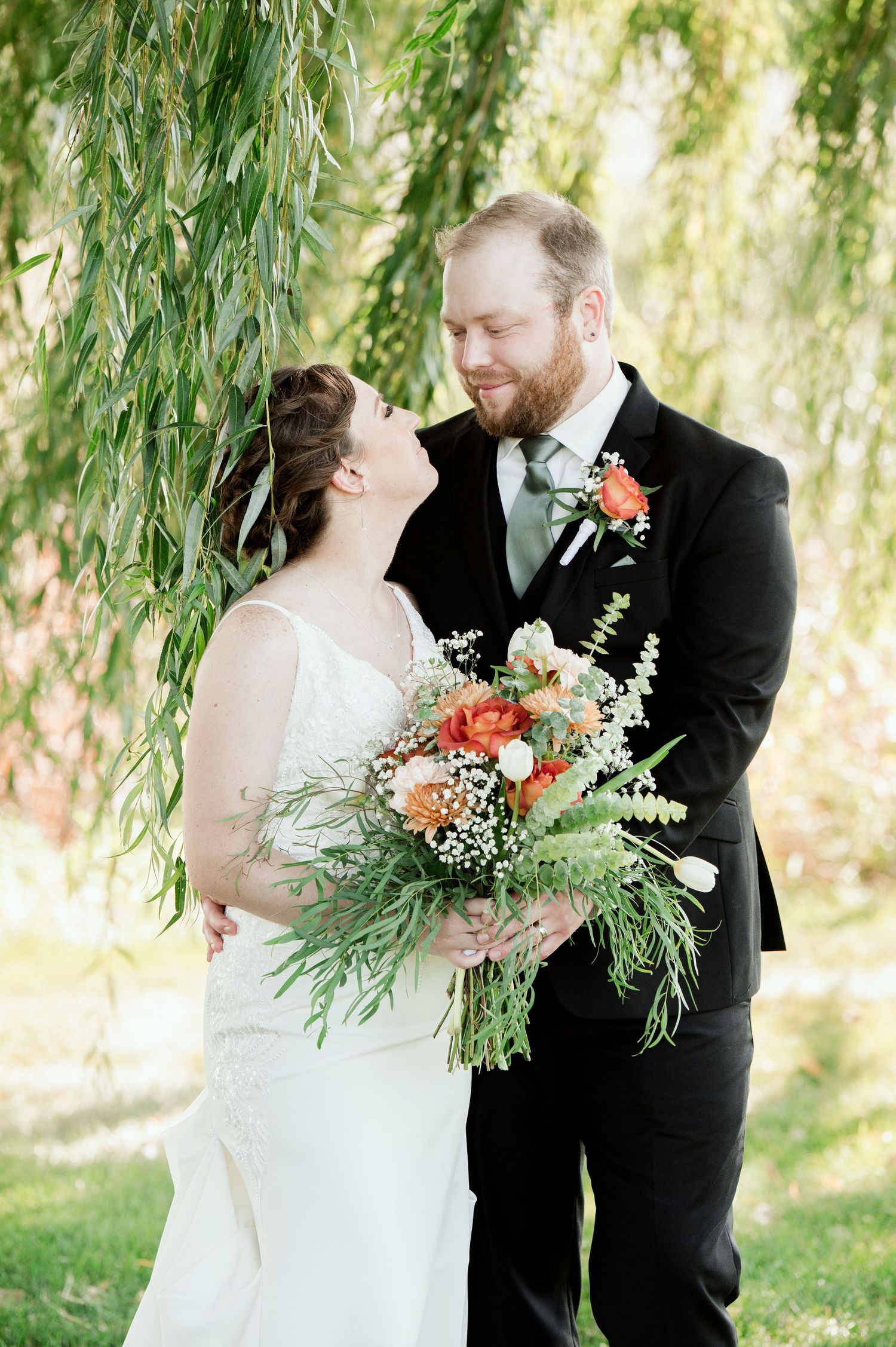 The Grands at Mulligans- Wedding Photos: couple stands beneath a weeping willow tree with a lush green and peach floral bouquet.