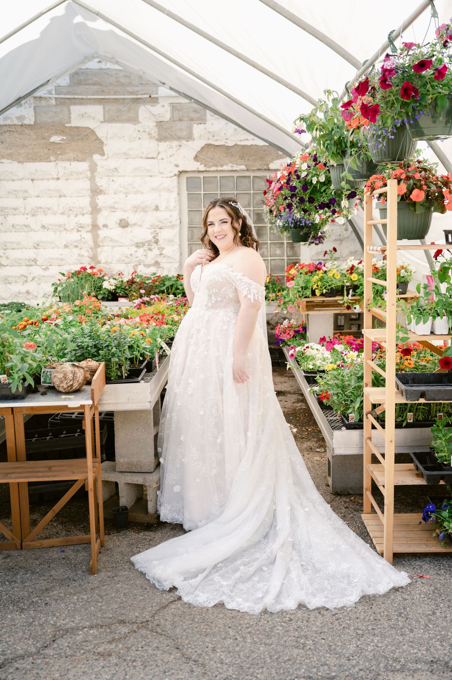 Elegant white wedding gown with long train photographed in a greenhouse filled with vibrant floral displays.