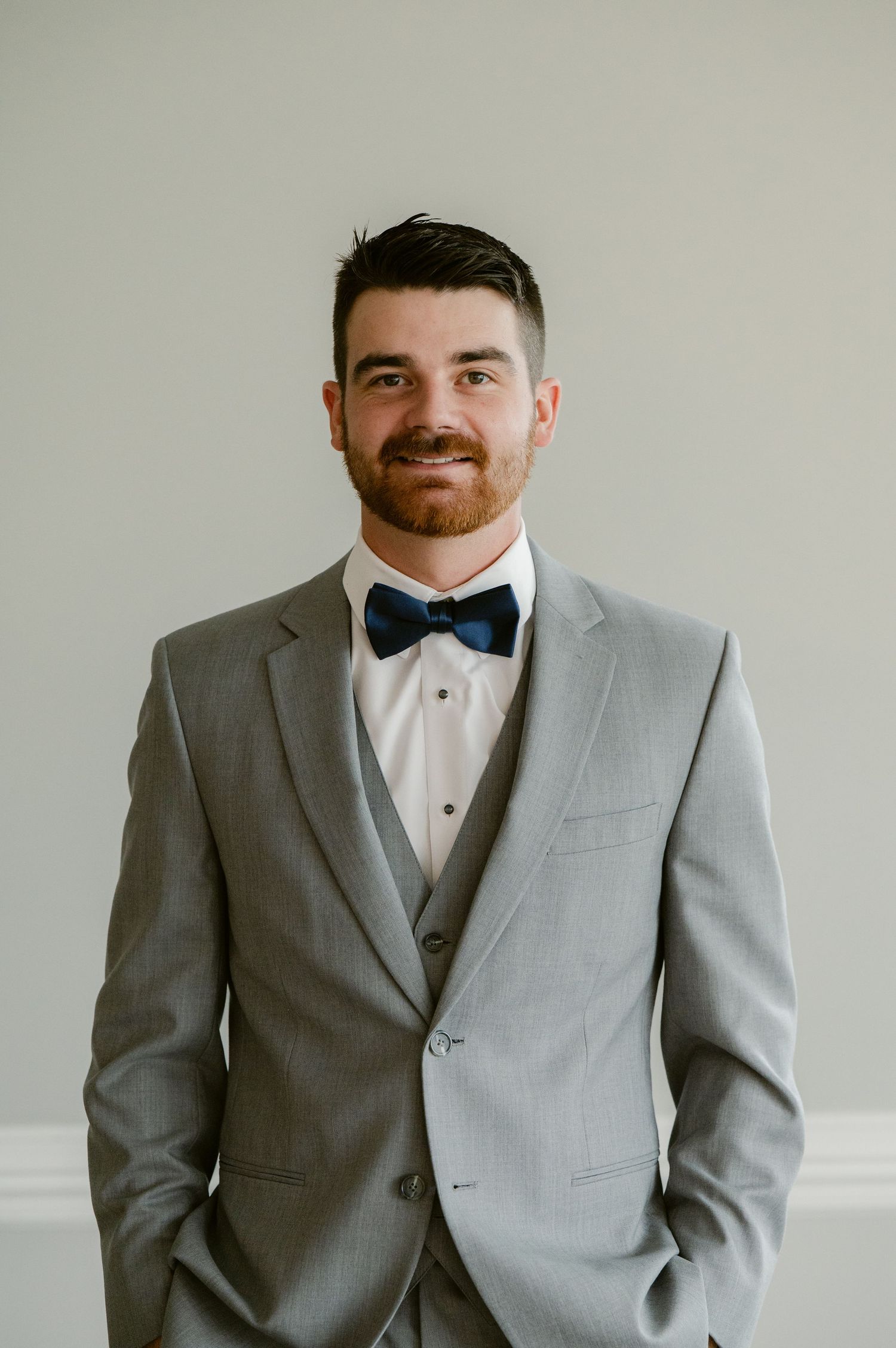 A person wearing a light grey three-piece suit with a navy blue bow tie poses against a neutral background.