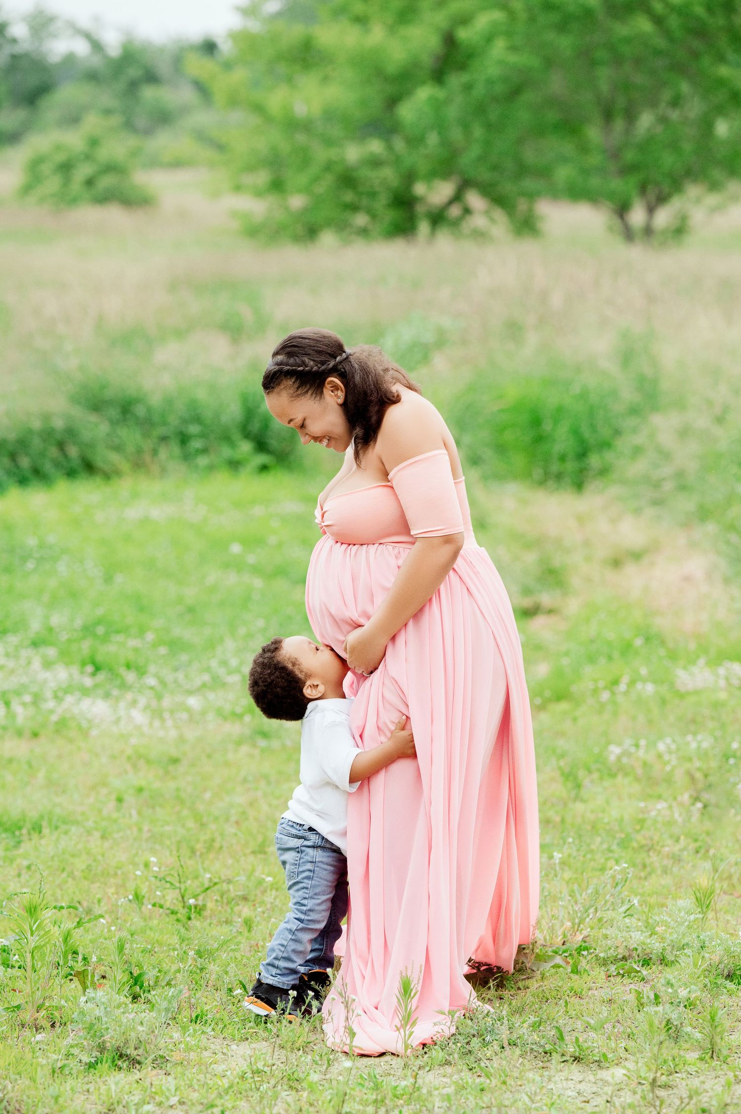 Pregnant mother in coral dress shares sweet moment with toddler in flowering meadow.