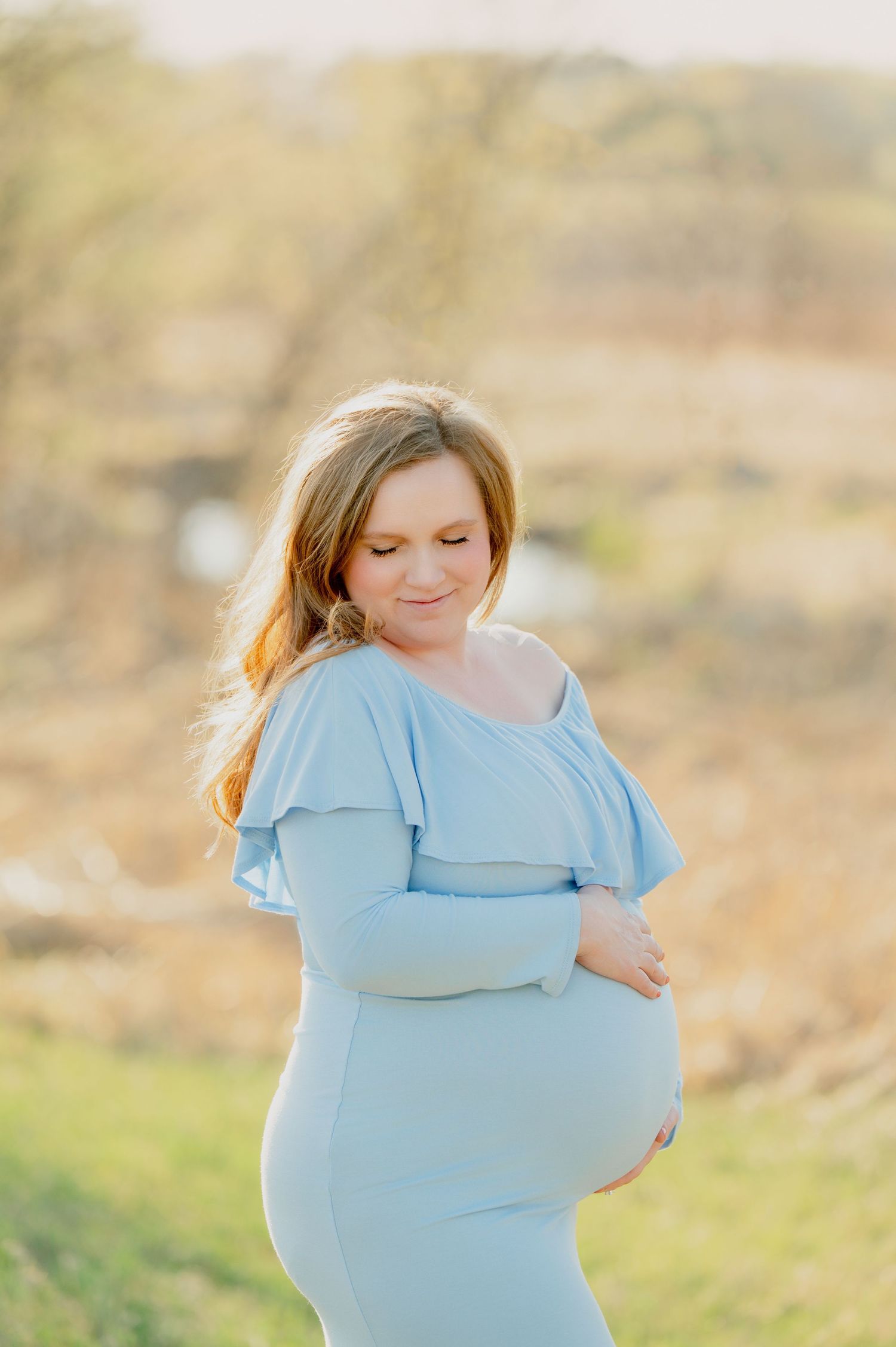 Expectant mother in flowing light blue dress poses in natural outdoor setting.