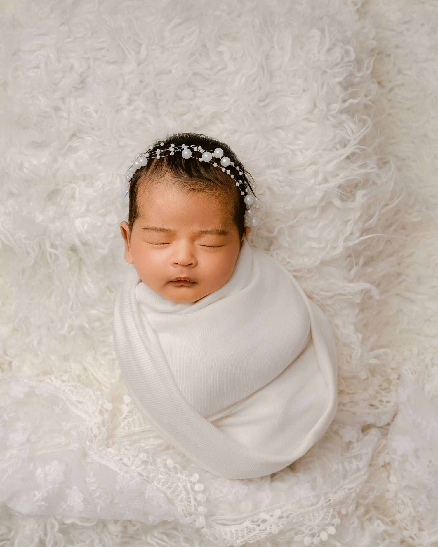 Newborn wrapped in white swaddle with pearl headpiece resting on textured white fur background.