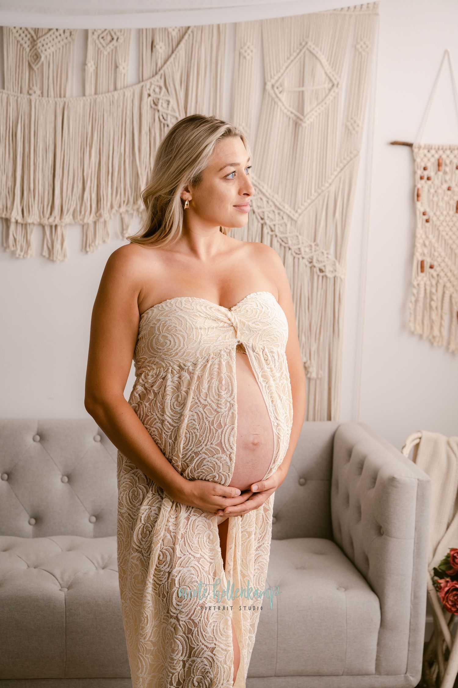 A pregnant woman in a cream lace dress stands in front of decorative macrame wall hangings and a gray couch.