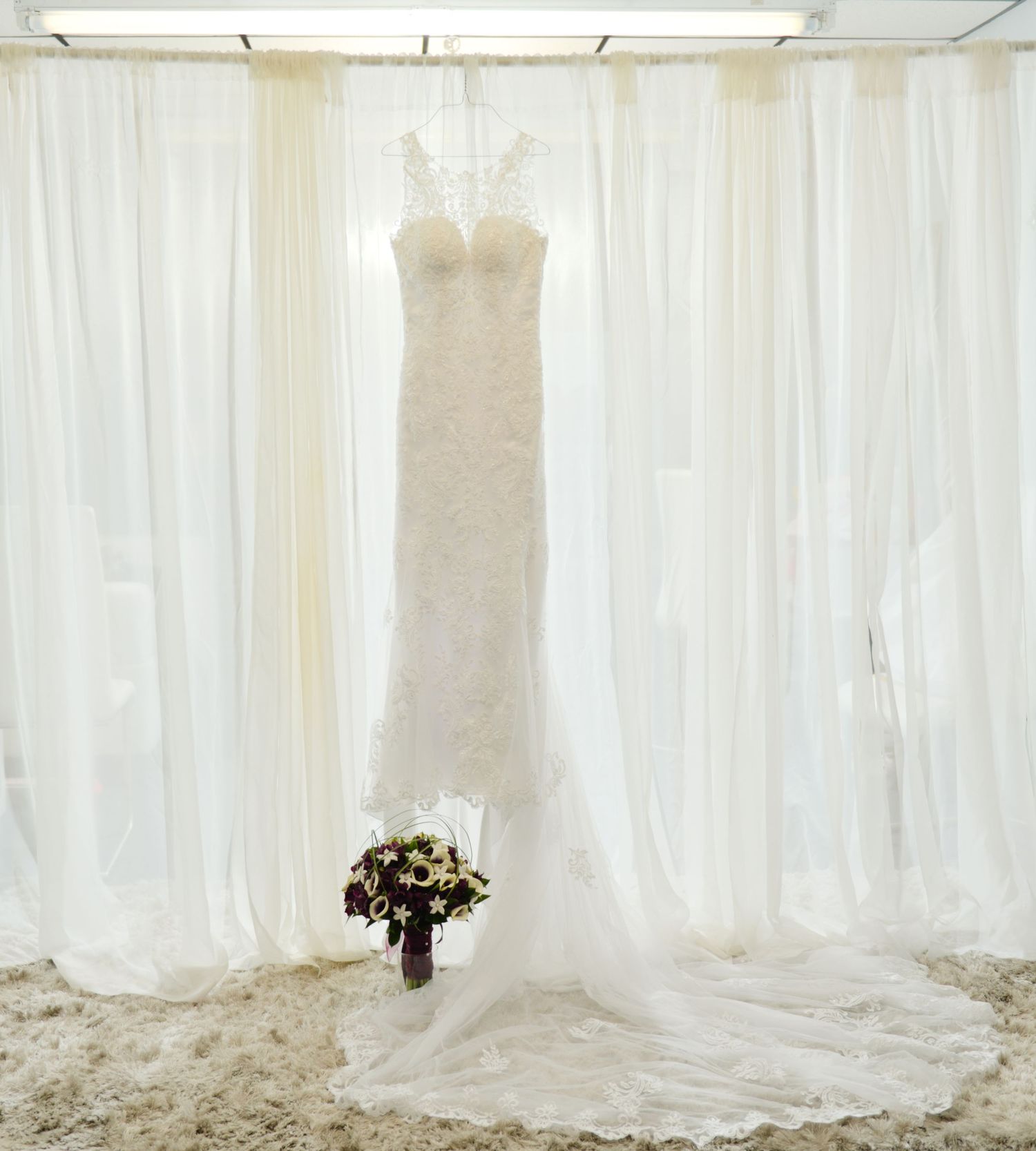 White lace wedding dress hanging against sheer white curtains with a dark bouquet at the bottom. Andover wedding at Magestic Oaks Golf Club