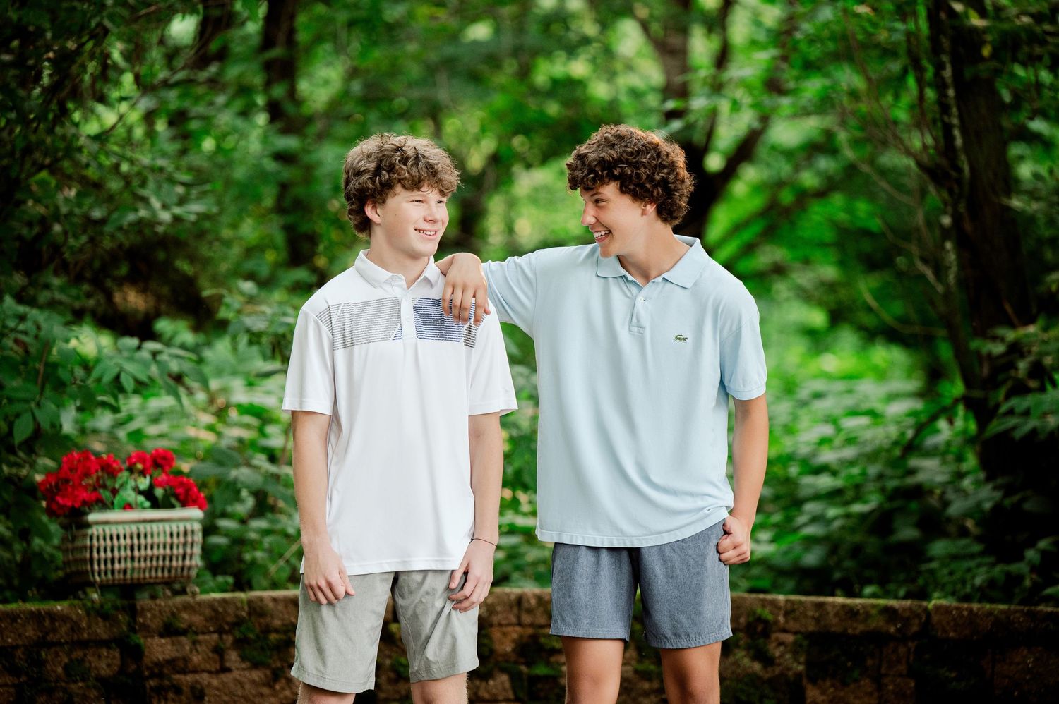 Clear Lake Family Photography: Two young people share a lighthearted moment on a stone wall with red flowers and forest greenery.