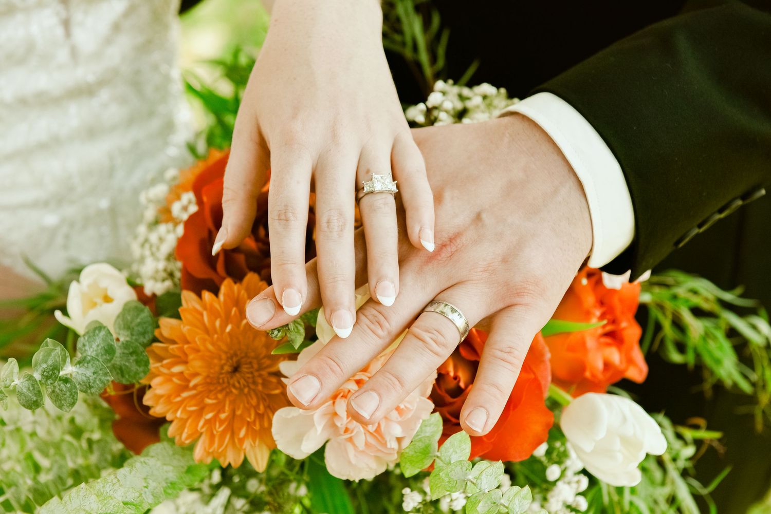 The Grands At Mulligans. Two hands with wedding rings displayed against colorful orange and white flowers in a bridal bouquet arrangement.