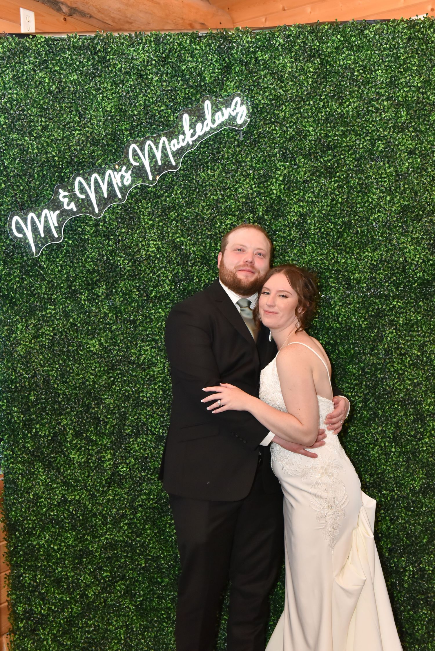 A couple embraces in front of a green hedge wall with 'Mr & Mrs' neon sign.