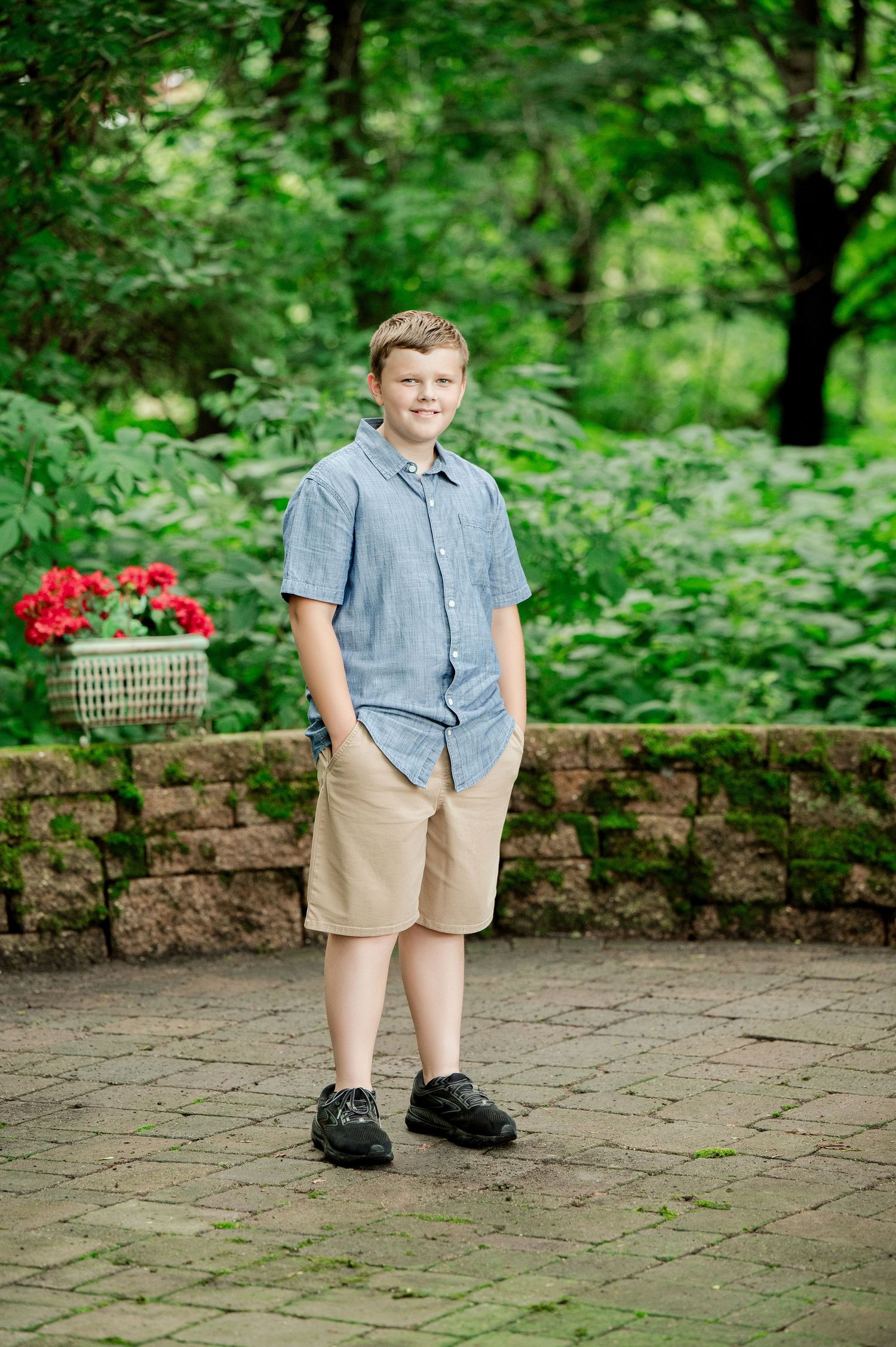 Young person stands on brick patio wearing blue button-down shirt and khaki shorts with lush green garden backdrop.