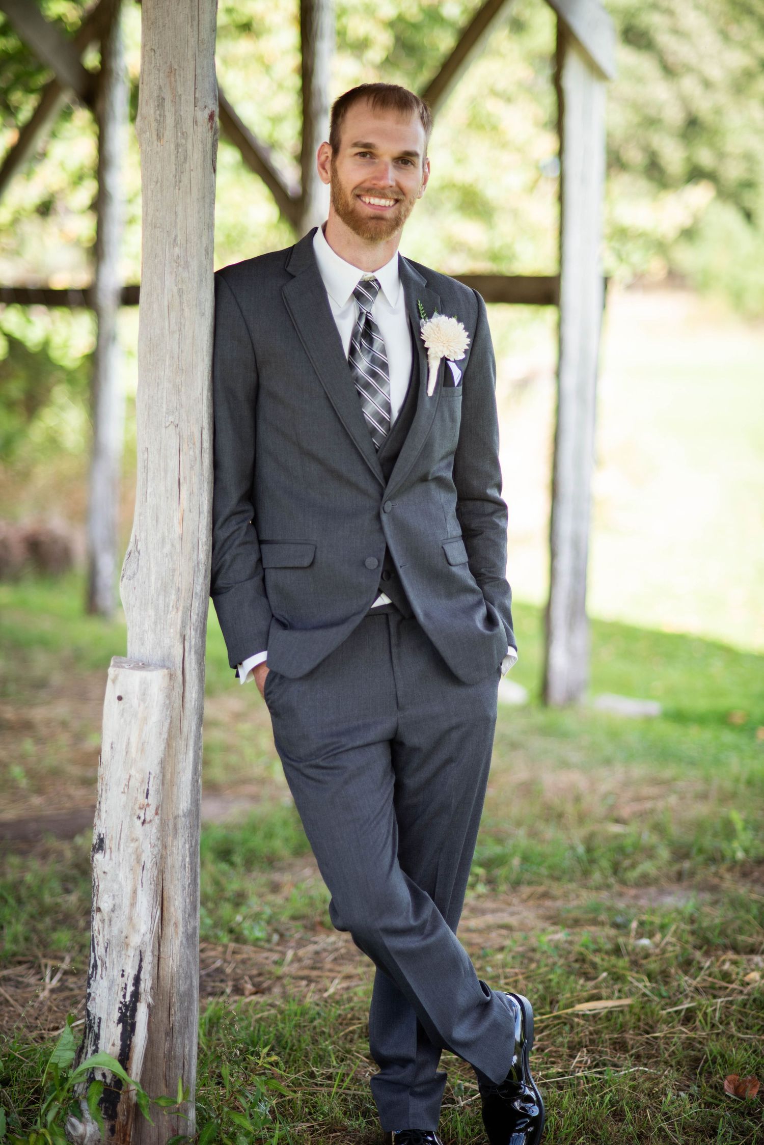Elegant groom in charcoal gray suit and striped tie poses casually against rustic white pillar outdoors.