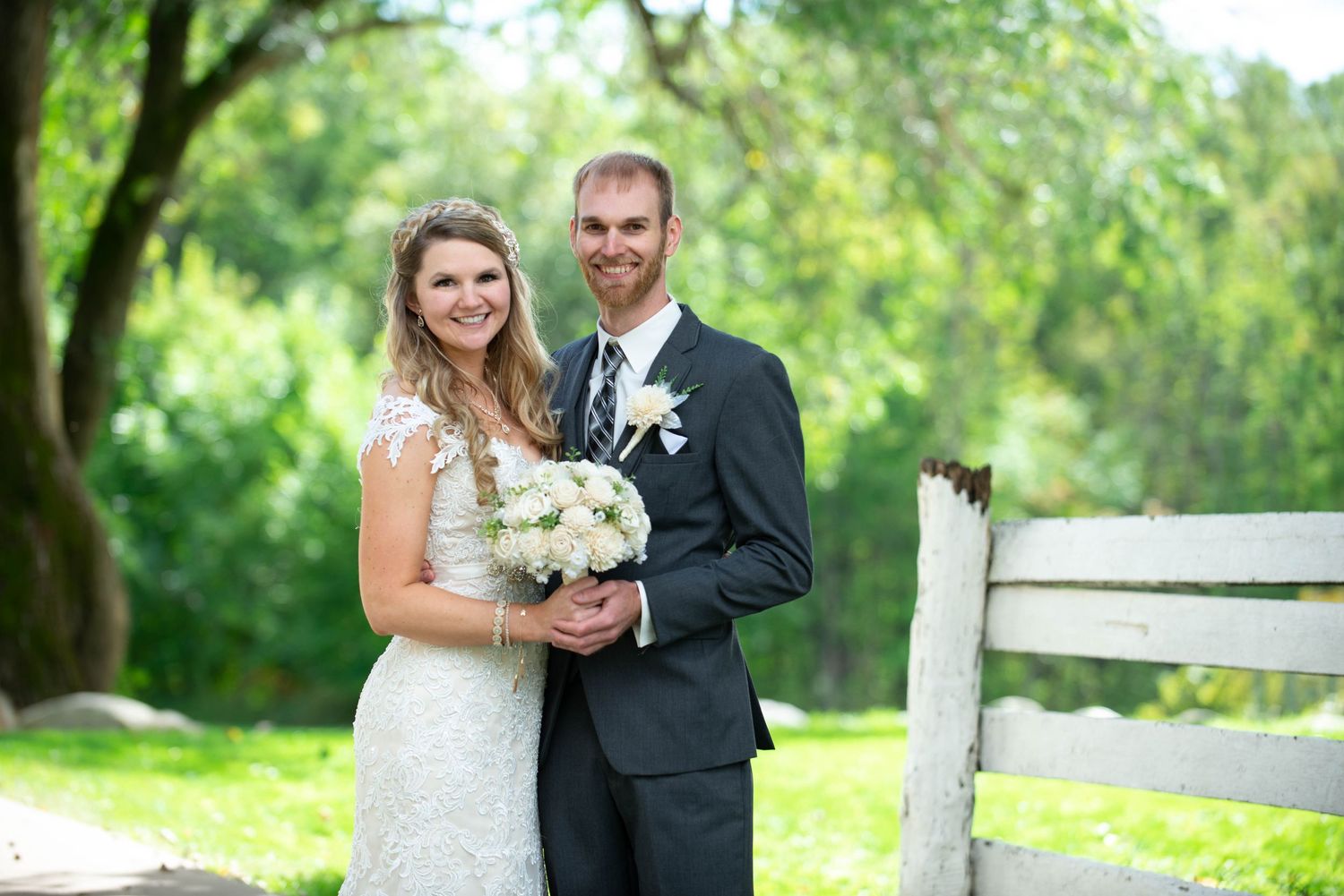 Newlyweds pose together by white fence on wedding day holding bouquet of white flowers. Rum River Barn Milaca Minnesota Weddings