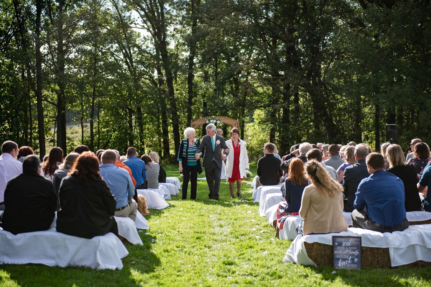 Outdoor wedding ceremony in a wooded setting with guests seated on white chairs on a grassy lawn. Rum River Barn Milaca Minnesota Weddings