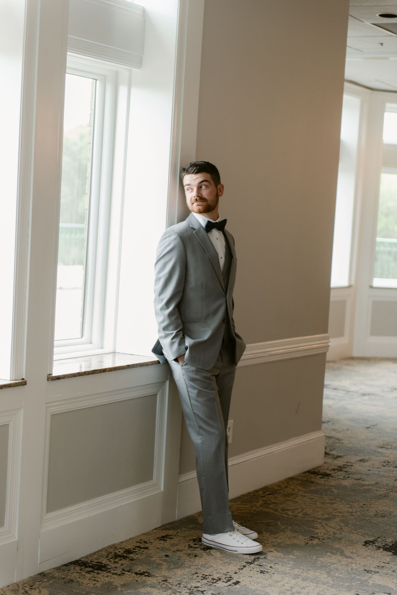 Person in a grey suit stands contemplatively beside bright windows in an empty interior space.