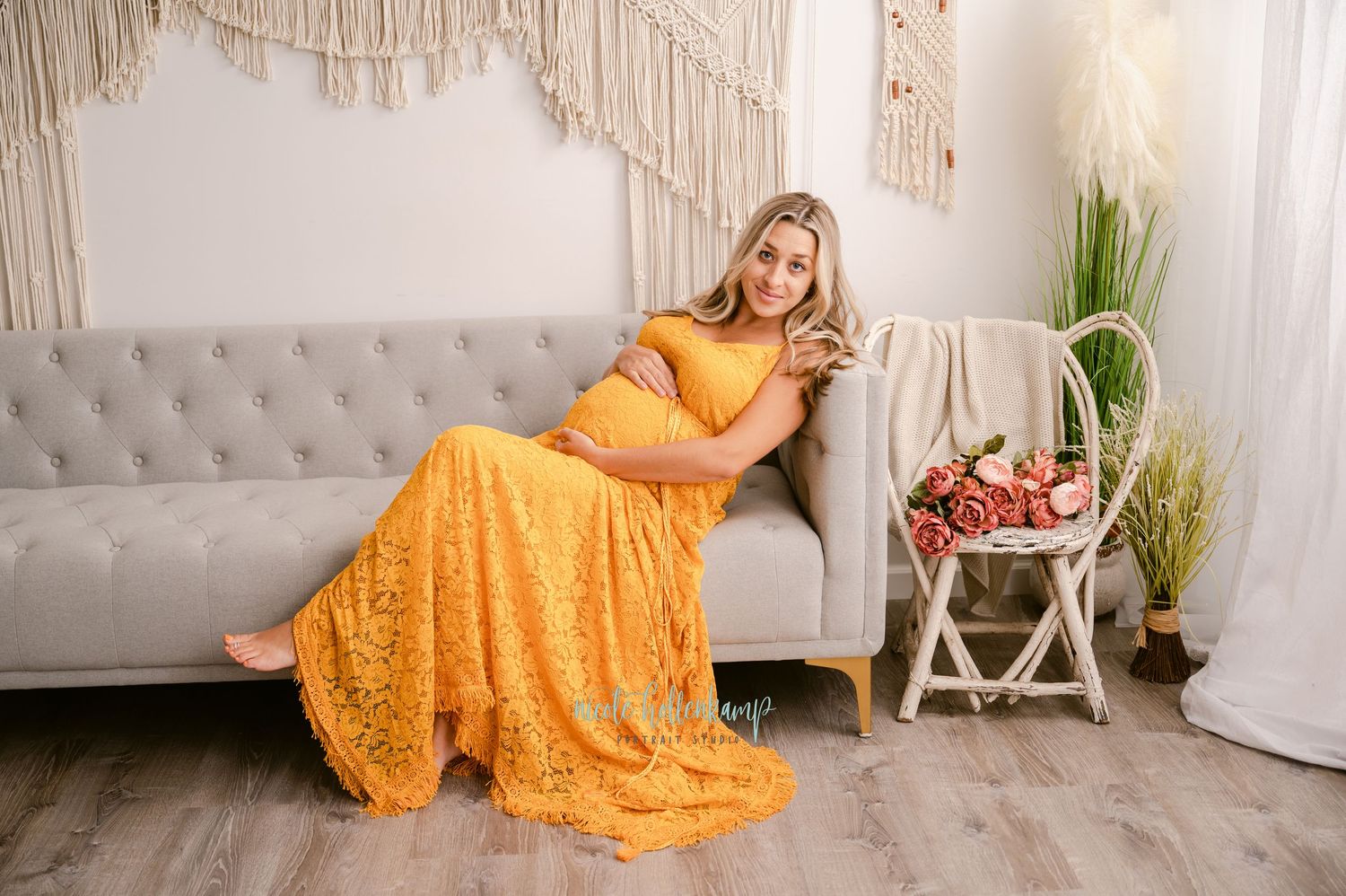 A figure in a flowing yellow lace gown reclines on a light gray tufted sofa next to macrame wall decor and plants.