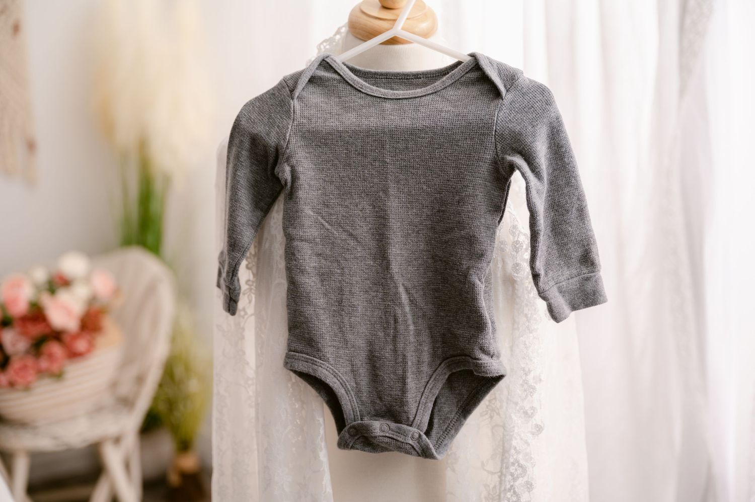 Gray long sleeve baby onesie hanging against white curtain with soft floral decor in background.